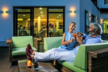 Paar auf Terrasse mit Drinks Ein Paar genießt Drinks auf einer Terrasse, umgeben von grünen Sofas. Im Hintergrund sind andere Gäste in einem Restaurant sichtbar.