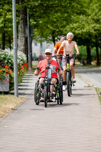 Drei Personen fahren auf einem Radweg. Eine Person in einem Handbike und zwei weitere auf Fahrrädern. Im Hintergrund ist ein Informationsschild und blühende Blumen sichtbar.