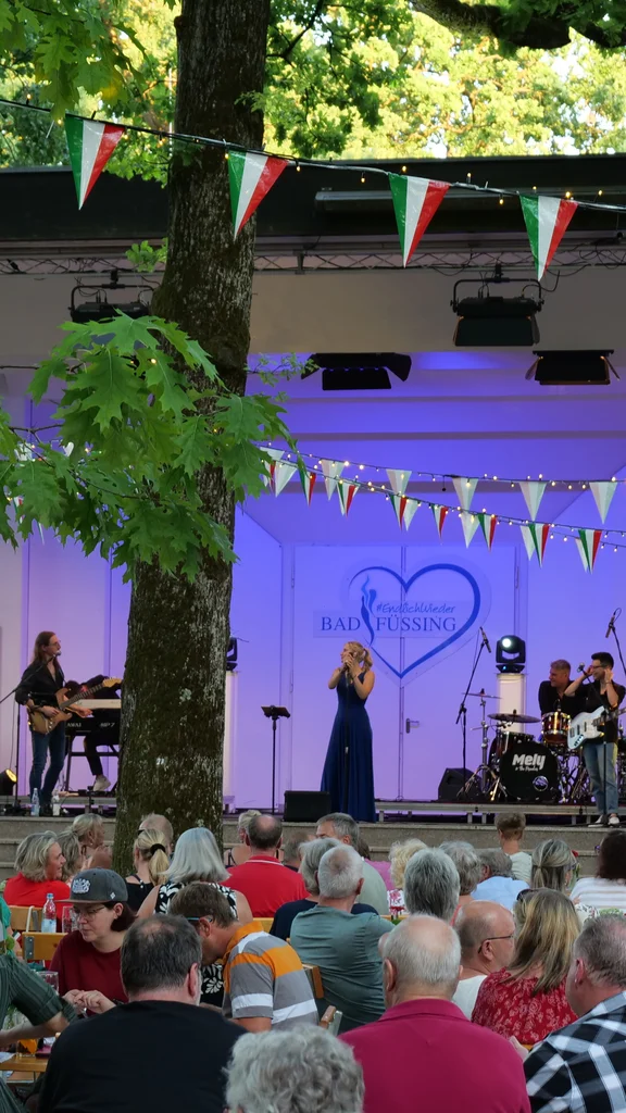 Ein Publikum sitzt an Tischen vor einer Freilichtbühne und sieht eine Liveband mit Sängerin, Gitarrist und Schlagzeuger unter bunten Wimpeln in einem Park.