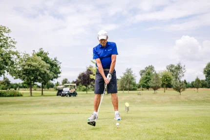 Mann schlägt Golfball auf Platz Ein älterer Mann in blauer Golfkleidung steht auf einem Golfplatz und bereitet sich darauf vor, einen Golfball zu schlagen. Im Hintergrund sind Bäume und ein Golfcart sichtbar.