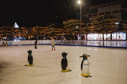 Nächtliche Eislaufbahn Nächtliche Eislaufbahn im Freien mit drei Pinguin-Lernhilfen im Vordergrund und zwei Personen auf dem Eis im Hintergrund.