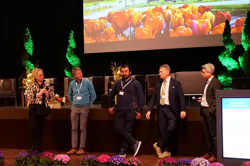 Conference Discussion on Stage A conference setting with five speakers on stage discussing. In the background, a large screen displays a scenic view of a park with tulips. Floral decorations are arranged on the stage and tables.