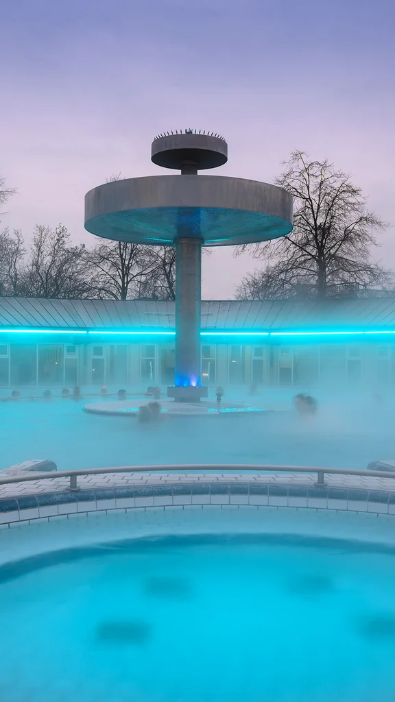 Dampfbad im Freien mit einem großen kreisförmigen Becken, mehreren badenden Personen und zentralem überdachten Brunnen oder Duschkonstruktion bei Dämmerung.