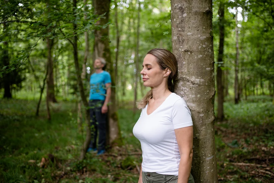 Eine Frau steht mit geschlossenen Augen an einem Baumstamm im Wald, während ein Mann weiter weg steht und den Blick nach oben richtet.