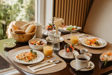 Ein Tisch mit einem üppigen Frühstück, einschließlich Rührei, frischem Obstsalat, Brot, Saft und Kaffee. Dekoratives Element ist eine kleine Vase mit Trockenblumen.