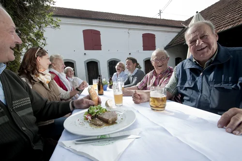 A group of older adults enjoying a meal outdoors at a long table, with plates of food and drinks in front of them, smiling and laughing together in a festive atmosphere.