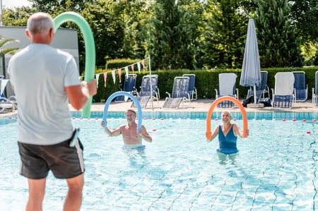 Schwimmtraining im Pool Eine Gruppe von Personen steht im Pool mit Aufblasringen. Der Trainer zeigt den Teilnehmern eine Übung. Sonnige Atmosphäre mit Liegen im Hintergrund.