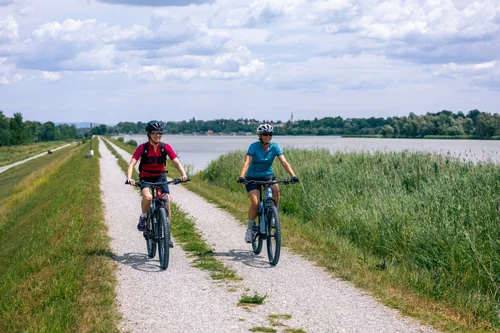 Frauen radeln am See Zwei Frauen fahren mit Fahrrädern auf einem Schotterweg entlang eines Sees, mit Gras und Schilf am Ufer und bewölktem Himmel.
