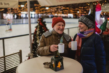 Frauen an der Eisbahn Zwei Frauen im Winterkleidung sitzen an einem Tisch vor einer Eisbahn und halten Tassen. Im Hintergrund sind Lichter und ein geschmückter Weihnachtsbaum zu sehen.
