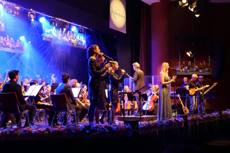 Eine Live-Bühne mit Sängern und einem Orchester. Vier Sänger in eleganten Kleidern stehen vorne, während ein Orchester im Hintergrund spielt. Lichtstrahlen und florale Dekorationen sind zu sehen.