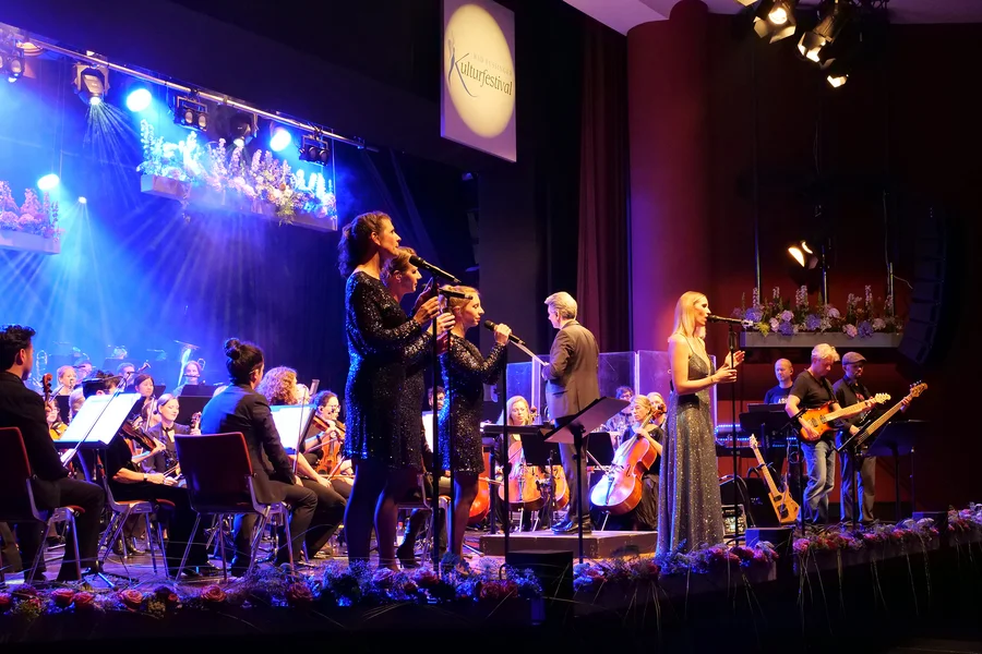 Eine Live-Bühne mit Sängern und einem Orchester. Vier Sänger in eleganten Kleidern stehen vorne, während ein Orchester im Hintergrund spielt. Lichtstrahlen und florale Dekorationen sind zu sehen.