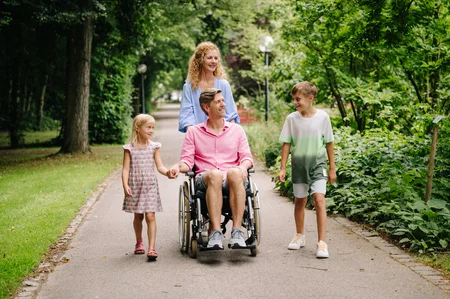 Eine Familie geht in einem Park. Ein Mann im Rollstuhl lächelt, während er von zwei Kindern und einer Frau begleitet wird. Die Kinder halten Hände und zeigen Freude. Umgebende Bäume und Sträucher schaffen eine grüne Atmosphäre.