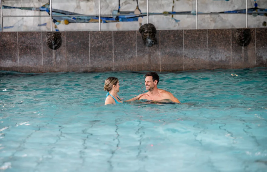 Paar im Schwimmbad Ein Mann und eine Frau stehen im Wasser eines Schwimmbads und unterhalten sich. Die Frau trägt einen Badeanzug, der Mann ist oberkörperfrei. Es gibt einen dekorativen Wasserfall im Hintergrund.