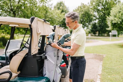 Mann packt Golfausrüstung aus Ein Mann mit Brille packt Golfschläger aus einem Golftaschen. Im Hintergrund sind andere Golfwagen und Bäume sichtbar.