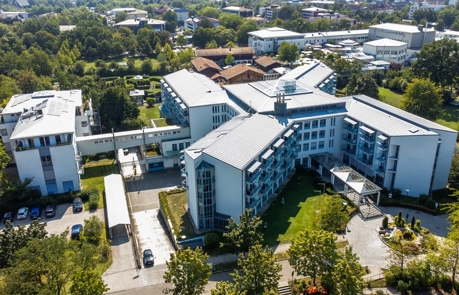Eine Luftaufnahme eines modernen Hotels mit angrenzenden Gärten und Parkplätzen. Mehrere Gebäude sind sichtbar, darunter ein Hauptgebäude mit großen Fenstern.