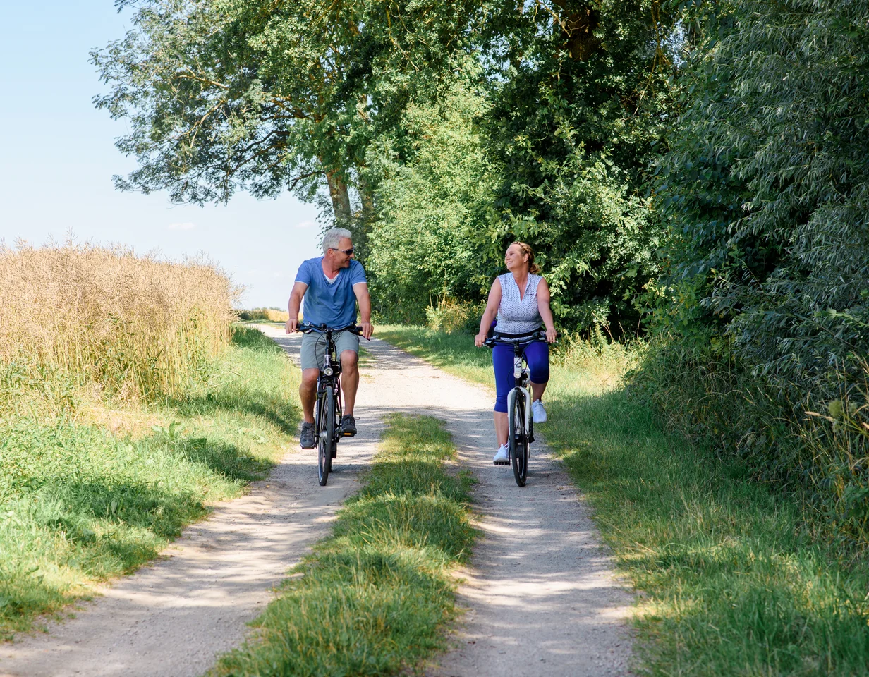 Ein Mann und eine Frau fahren mit Fahrrädern auf einem Schotterweg, umgeben von grünen Bäumen und Getreidefeldern. Es ist sonnig und die Stimmung ist entspannt, während sie miteinander sprechen.
