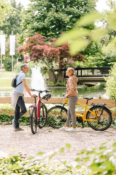 Ein Mann und eine Frau stehen mit Fahrrädern am Rand eines Parks, während im Hintergrund Senioren auf Bänken sitzen. Ein Wasserspiel bricht die ruhige Szene.