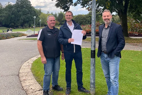 Drei Männer im Park Drei Männer stehen in einem Park neben einer Straßenlaterne; der Mann in der Mitte hält ein Dokument in der Hand.