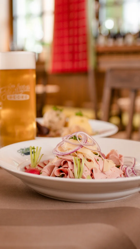 Ein gedeckter Tisch mit Biergläsern, Schinken auf einem Teller, einer Schüssel mit Soße und frischem Brot. Der Hintergrund zeigt eine gemütliche, rustikale Umgebung.