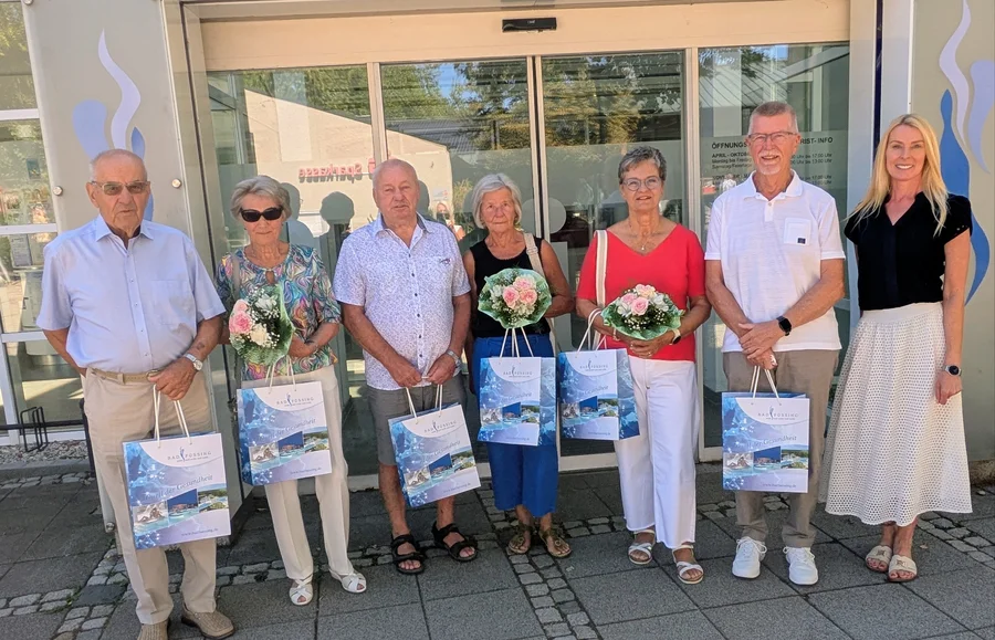 Gruppenszene vor dem Eingang, sechs Personen halten Blumensträuße und Tüten vor einer Glasfront. Alle lächeln freundlich in die Kamera.