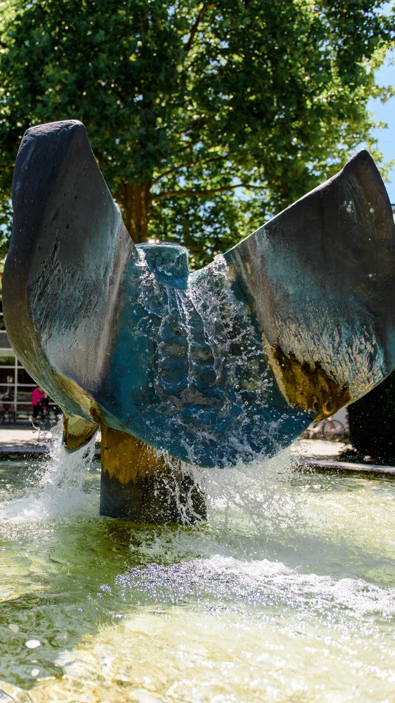Ein Springbrunnen mit einer großen, stilisierten Skulptur in der Mitte, umgeben von klarem Wasser. Im Hintergrund sieht man ein modernes Gebäude und einige Bäume. Menschen radeln in der Nähe.