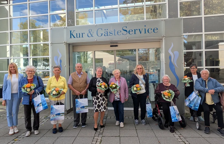 Gruppe vor dem Kurservice Vor einem Gebäude mit der Aufschrift „Kur & GästeService“ stehen mehrere ältere Personen nebeneinander, einige halten Blumensträuße und Geschenktüten; zwei Personen sitzen auf Rollatoren.