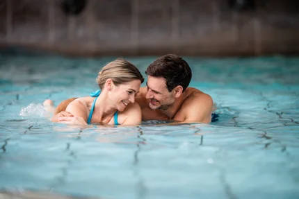 Ein Paar lächelt und spielt im Wasser eines Schwimmbads. Beide haben einen entspannten Gesichtsausdruck. Das Wasser ist klar und reflektiert Licht.