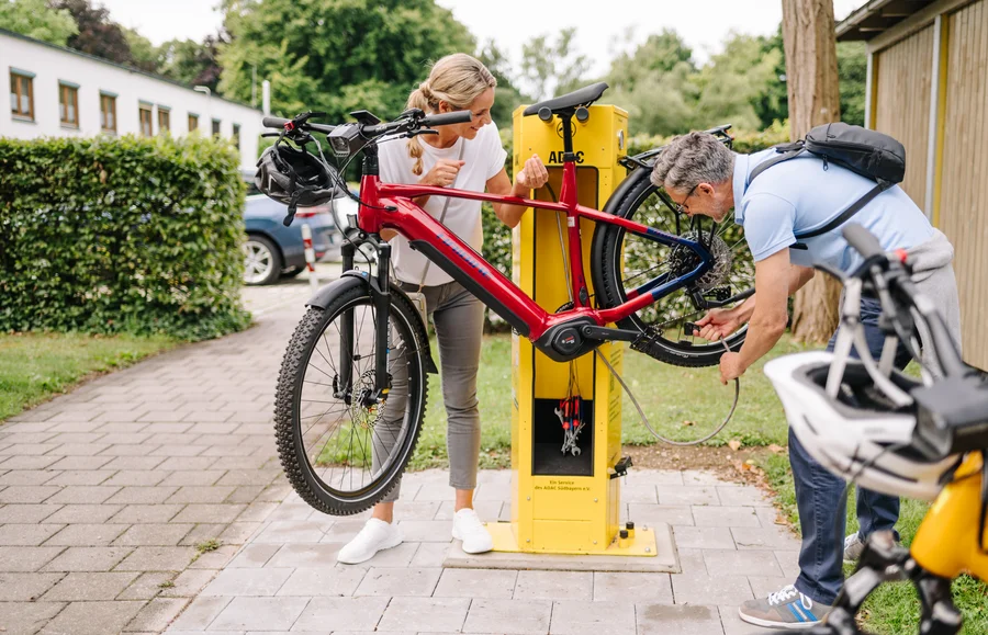 Eine Frau und ein Mann reparieren gemeinsam ein rotes Elektrofahrrad an einer gelben Fahrradservicestation im Freien.