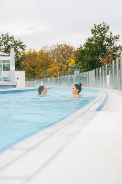 Zwei Frauen entspannen im warmen Wasser eines Pools, umgeben von herbstlichen Bäumen im Hintergrund. Es ist eine ruhige, entspannende Atmosphäre mit leichtem Dampf über dem Wasser.