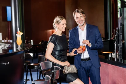 Paar in einer eleganten Bar Ein Mann und eine Frau stehen in einer Bar. Der Mann trägt einen Anzug und hält ein Getränk, während die Frau in einem glitzernden Kleid sitzt und eine Clutch trägt. Sie lächeln freundlich.