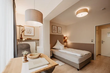 Ein helles Hotelzimmer mit einem Einzelbett, Holzarbeitsplatz, Tisch, Lampen und dekorativen Bildern an der Wand.