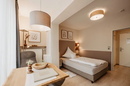 Ein helles Hotelzimmer mit einem Einzelbett, Holzarbeitsplatz, Tisch, Lampen und dekorativen Bildern an der Wand.
