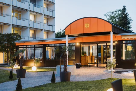 Ein modernes Hotelgebäude mit orangefarbenem Dach und verglasten Eingangsbereichen. Umgeben von Pflanzen und einer gepflegten Gartenlandschaft, mit runden Lichtern und kleinen Bäumen auf einem gepflasterten Weg.