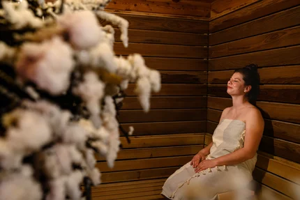 Frau in der Sauna Eine Frau sitzt mit geschlossenen Augen auf einer Holzbank in einer Sauna, in ein Handtuch gewickelt. Im Vordergrund ist verschwommen ein Bündel Sauna-Zweige zu sehen.