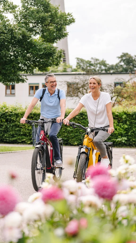 Zwei Personen radeln fröhlich auf einer Straße, umgeben von Blumen und grünen Büschen, mit Gebäuden im Hintergrund.