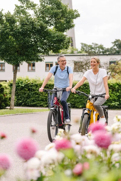 Zwei Radfahrer im Blumenbeet Zwei Personen radeln fröhlich auf einer Straße, umgeben von Blumen und grünen Büschen, mit Gebäuden im Hintergrund.