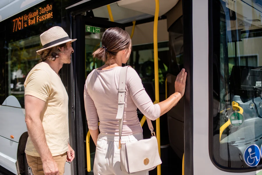 Warten vor dem Bus 776 Ein Mann und eine Frau stehen an einem Bus, während die Frau die Tür berührt und ein Schild zeigt. Die Busnummer ist 776 und zeigt Richtung Bad Füssing.