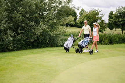 Paar mit Golfwagen auf Golfplatz Ein Mann und eine Frau stehen auf einem Golfplatz, beide halten Golfwagen mit Schlägern. Im Hintergrund sind Bäume und Gräser zu sehen.
