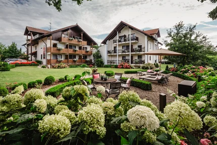 Gartenansicht eines Hotels Eine Unterkunft mit zwei Gebäuden umgeben von grünem Rasen und blühenden Blumen. Im Vordergrund befinden sich ein Gartentisch und Liegen unter einem Sonnenschirm.