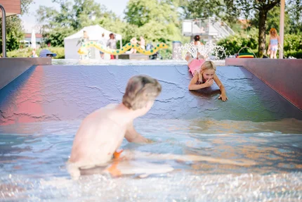Ein Kind rutscht in einem Wasserspielbereich, während ein Erwachsener im Wasser ist. Im Hintergrund sind weitere Kinder und Spielbereiche sichtbar.