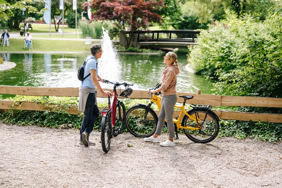 Ein Mann und eine Frau stehen mit Fahrrädern an einem Teich. Der Mann hat ein rot-schwarzes Fahrrad, während die Frau ein gelbes Fahrrad hat. Im Hintergrund sind Bäume und ein Springbrunnen zu sehen.