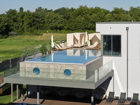 Rooftop Infinity Pool and Lounge A modern building features a rooftop infinity pool, surrounded by glass walls and sun loungers. Below, patio seating is visible, alongside a landscaped area with greenery.