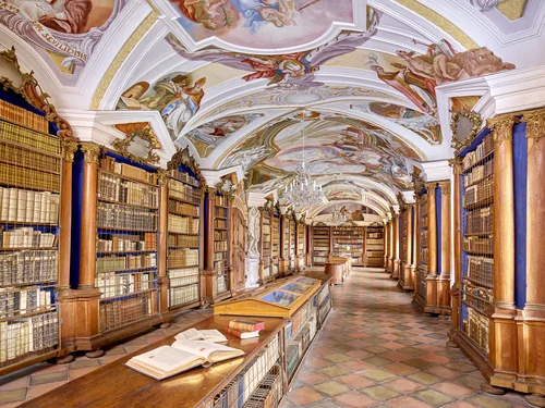 Eine Innenansicht einer bibliotheksähnlichen Halle mit hohen Bücherregalen, kunstvoll bemalten Decken und einem großen Kronleuchter in der Mitte. Vorne liegt ein offenes Buch auf einem Holzgestell.