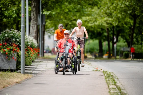 Drei Personen fahren mit Fahrrädern auf einem Bürgersteig, eine Person im Rollstuhl am vorderen Ende. Es sind Blumenbeete und Bäume im Hintergrund zu sehen.