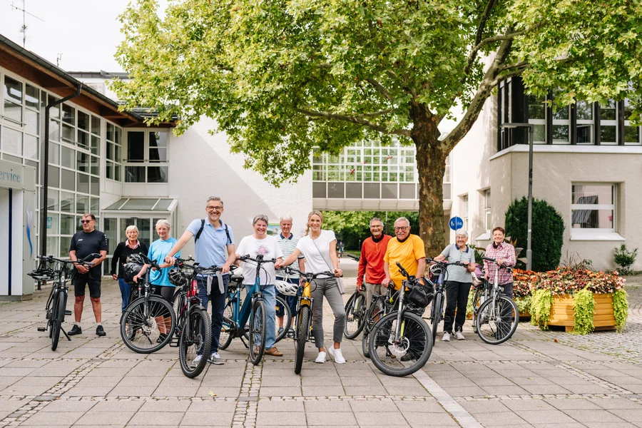 Eine Gruppe von Fahrradfahrern steht vor einem Gebäude, lächelnd und bereit für eine Ausfahrt. Sie haben verschiedene Fahrräder und tragen Sportbekleidung, umgeben von Pflanzen und Bäumen.