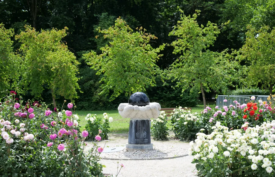 Eine Statue in einem Park, umgeben von bunten Blumenbeeten. Im Hintergrund sind grüne Bäume zu sehen.