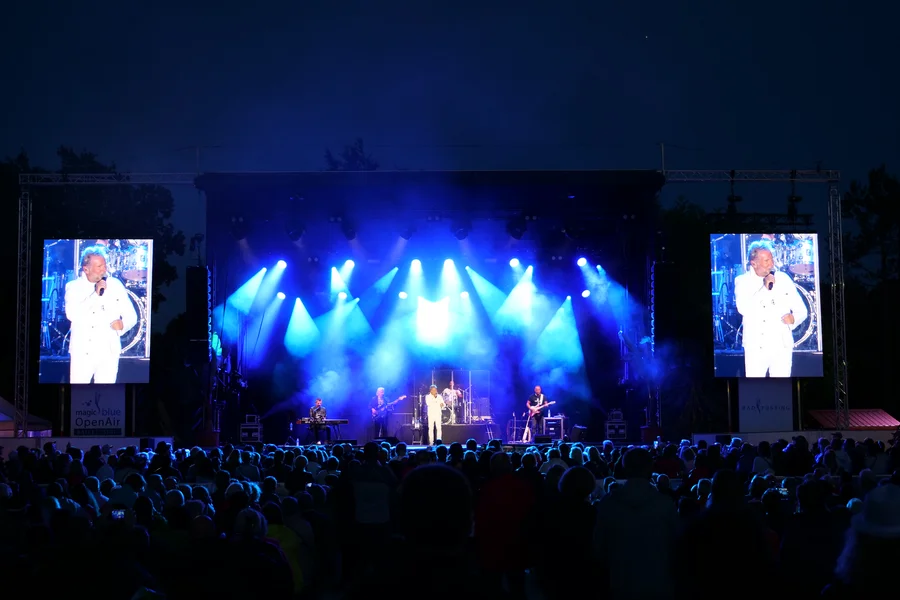 Ein Sänger in weißem Outfit performt auf einer Bühne mit mehreren Musikern, während das Publikum begeistert zuschaut. Großbildschirme zeigen die Darbietung, und die Bühne ist mit blauen Lichtern beleuchtet.