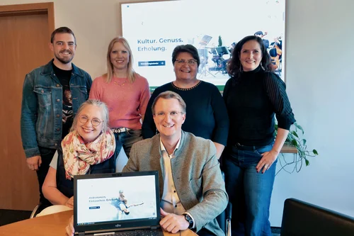 Gruppenfoto im Büro Sechs Personen posieren in einem Büro; vorne sitzt ein Mann mit einem Laptop, auf dem eine Webseite zu sehen ist. Im Hintergrund stehen vier weitere Personen vor einem Bildschirm mit der Aufschrift „Kultur. Genuss. Erholung.“