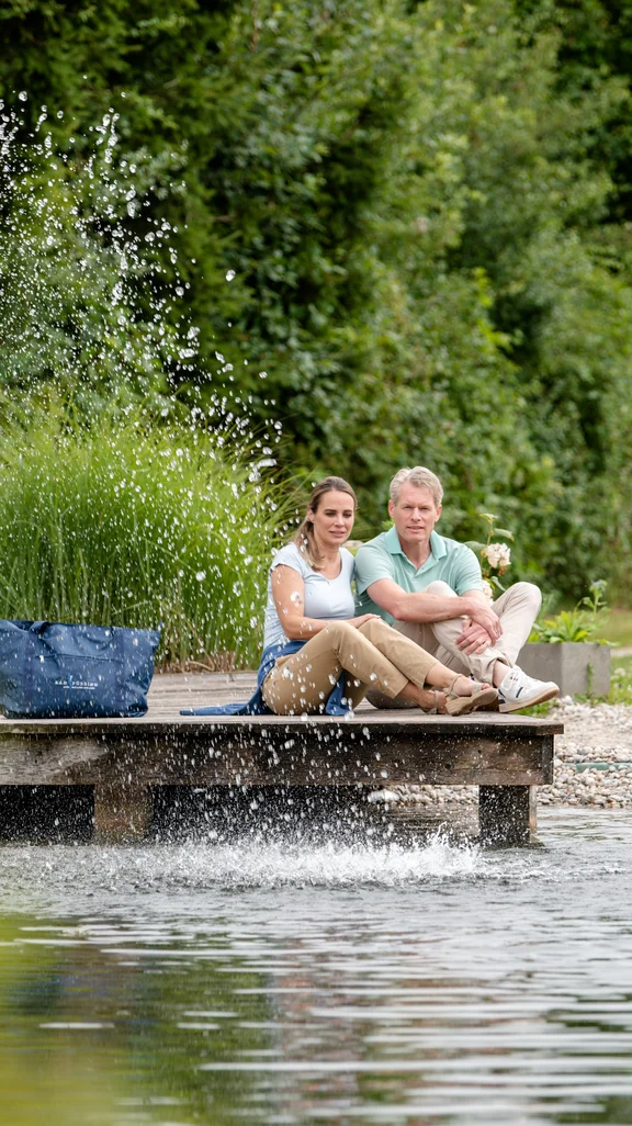 Ein Paar sitzt auf einem Holzsteg am Wasser. Im Hintergrund ist ein Wohnmobil zu sehen und es spritzt Wasser aus dem Teich. Grüne Pflanzen umgeben die Szene.
