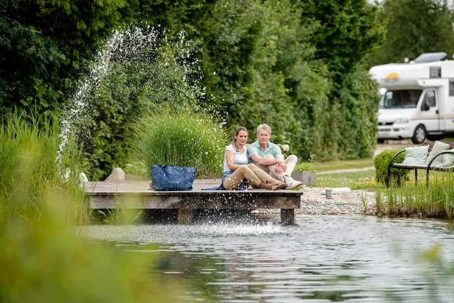 Paar am Teich mit Wohnmobil Ein Paar sitzt auf einem Holzsteg am Wasser. Im Hintergrund ist ein Wohnmobil zu sehen und es spritzt Wasser aus dem Teich. Grüne Pflanzen umgeben die Szene.